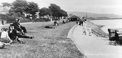 West Esplanade
Relaxing on the grass of the West Esplanade in Helensburgh, with big trees in the gardens of the seafront villas and one of the now demolished shelters in the distance. Image circa 1935.
